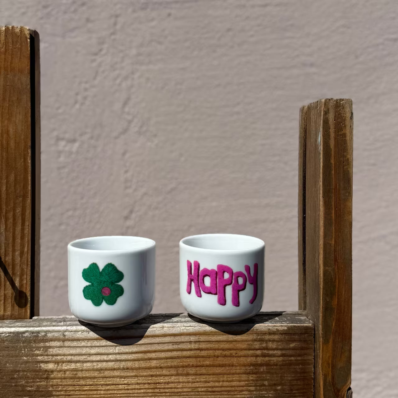 Zwei weiße Espressobecher mit Kleeblatt und violettem Happy Print stehen auf Holzgestell