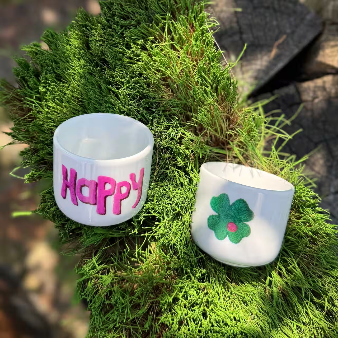 Zwei weiße Espressobecher mit Kleeblatt und violettem Happy Print liegen auf grünen Borsten