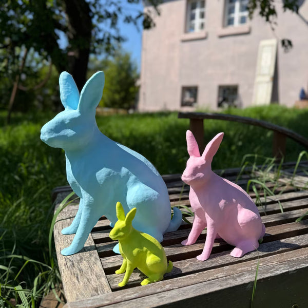 Ein blauer, rosa und limettengrüner 3D-gedruckter Hase sitzen auf Holzliege im Gras