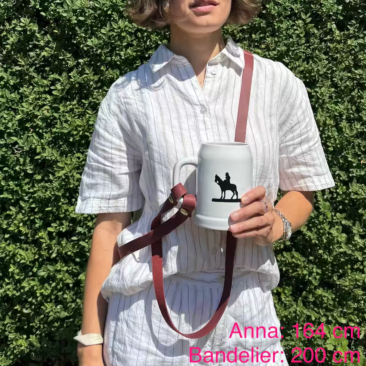 Model mit weißem Krug und rotem Lederband umgehängt steht in der Natur. Beschriftet mit "Anna: 164 cm, Bandelier: 200 cm"