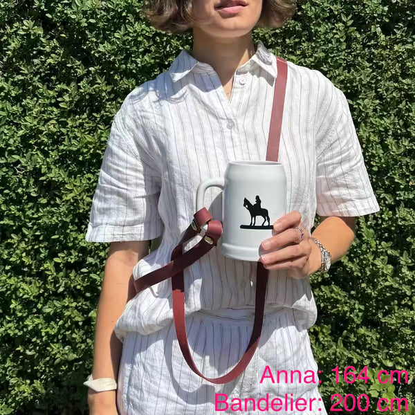 Model mit weißem Krug und rotem Lederband umgehängt steht in der Natur. Beschriftet mit "Anna: 164 cm, Bandelier: 200 cm"
