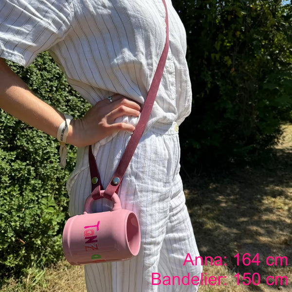 Model mit rosa Krug und magenta Lederband umgehängt steht in der Natur. Beschriftet mit "Anna: 164 cm, Bandelier: 150 cm"