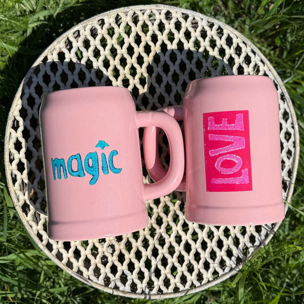 Zwei rosa Bierkrüge mit love und magic Print liegen auf Gitter im Gras