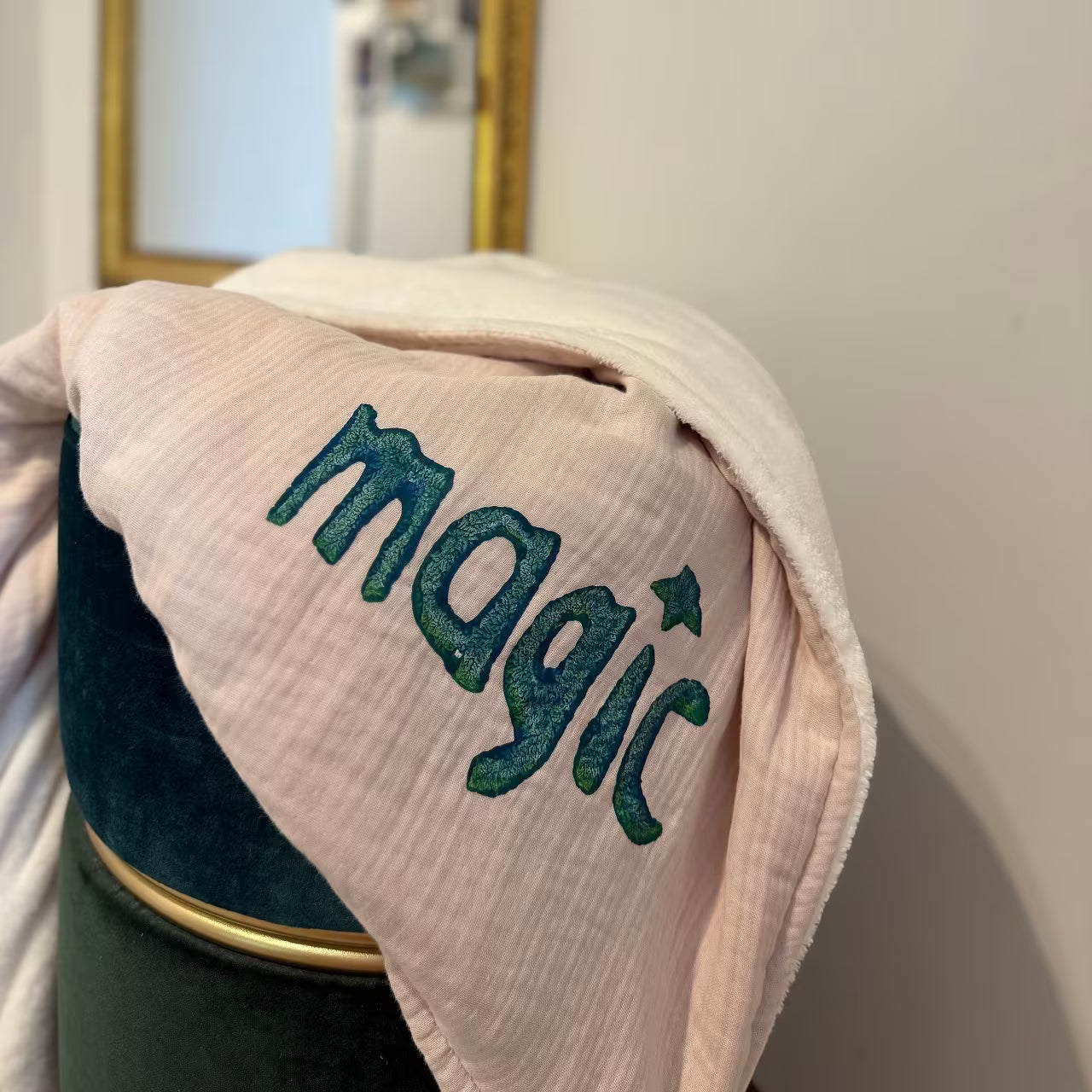 Leichtrosa Musselin Decke mit weißem Fleece auf Rückseite, mit petrolem Print "magic" liegt über grünem Hocker vor goldenem Spiegel