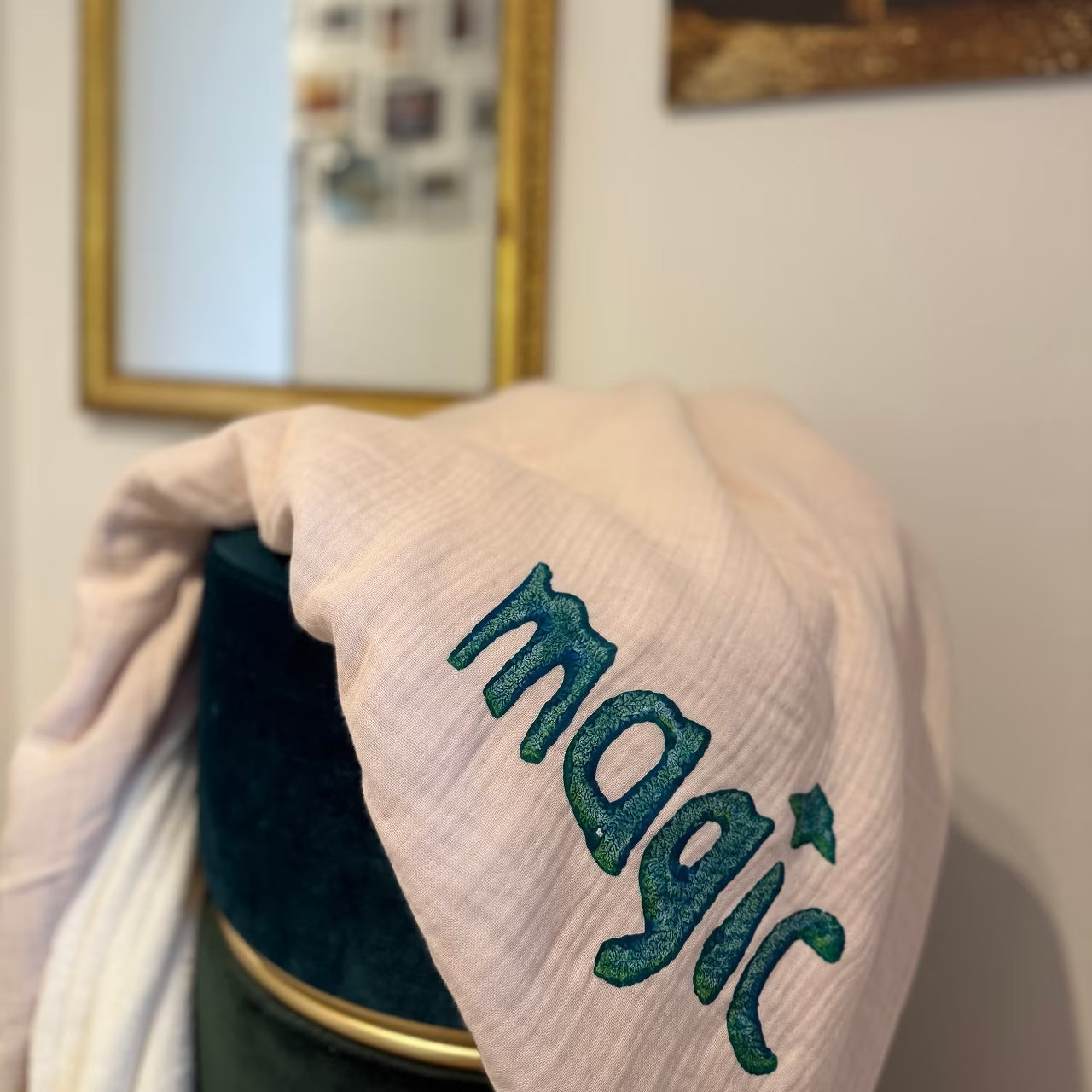 Leichtrosa Musselin Decke mit weißem Fleece auf Rückseite, mit petrolem Print "magic" liegt über grünem Hocker vor goldenem Spiegel