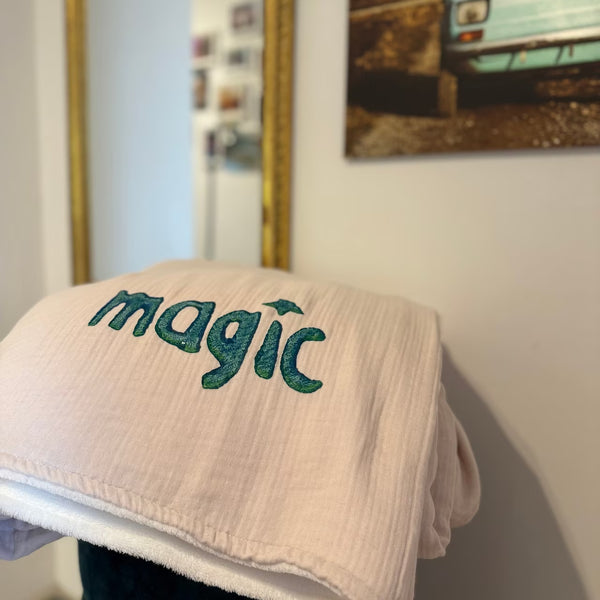 Leichtrosa Musselin Decke mit weißem Fleece auf Rückseite, mit petrolem Print "magic" liegt über grünem Hocker vor goldenem Spiegel und Fotografie