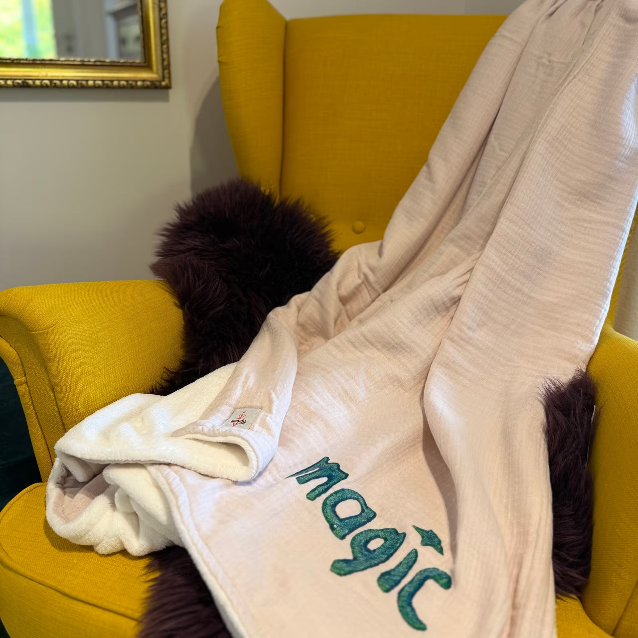 Leichtrosa Musselin Decke mit weißem Fleece auf Rückseite, mit petrolem Print "magic" liegt über senfgelbem Sessel und lila Fell