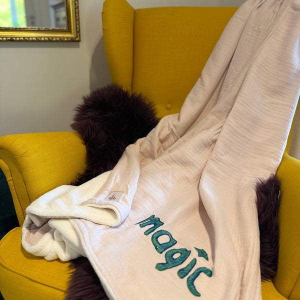 Leichtrosa Musselin Decke mit weißem Fleece auf Rückseite, mit petrolem Print "magic" liegt über senfgelbem Sessel und lila Fell