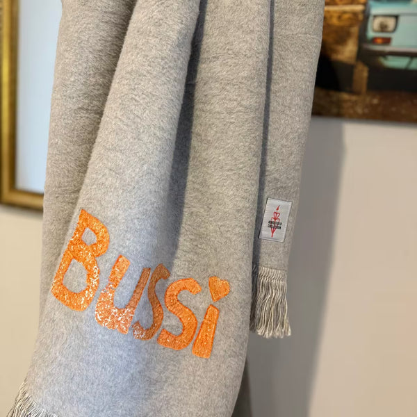 Graue Decke mit orangem Print "Bussi" hängt vor einem goldenen Spiegel und einer Fotografie