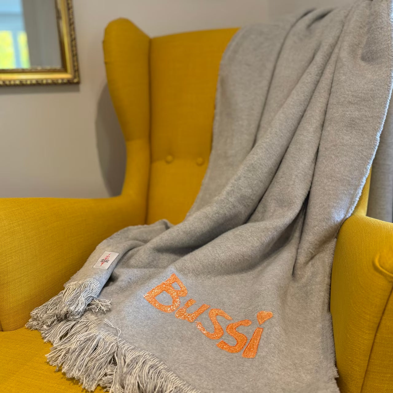 Graue Decke mit orangem Print "Bussi" liegt über senfgelbem Sessel vor goldenem Spiegel