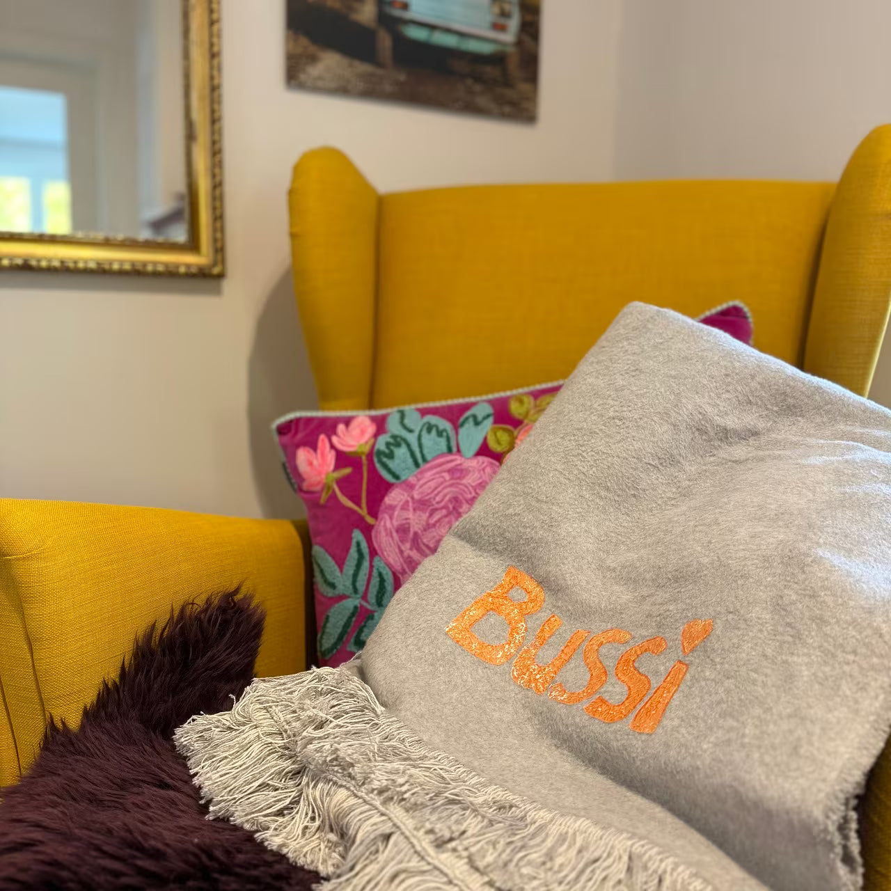 Graue Decke mit orangem Print "Bussi" liegt auf senfgelbem Sessel, lila Fell und buntem Kissen vor goldenem Spiegel und Fotografie