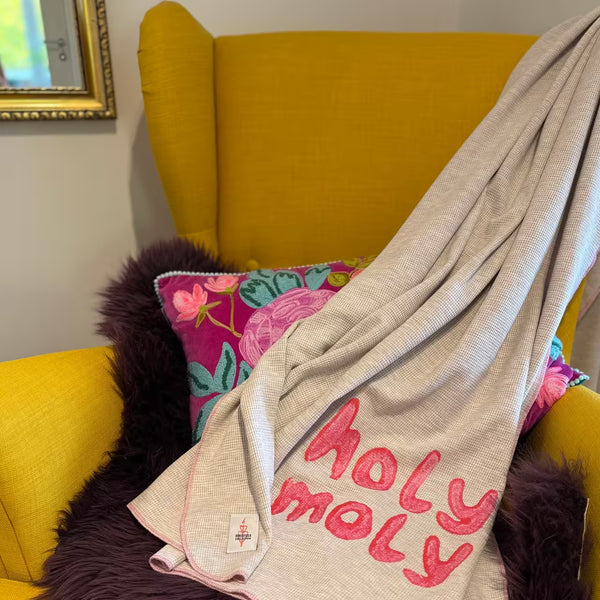 Graue leichte Decke mit pinkem Print "holy moly" liegt auf senfgelbem Sessel mit lila Fell und buntem Kissen vor goldenem Spiegel