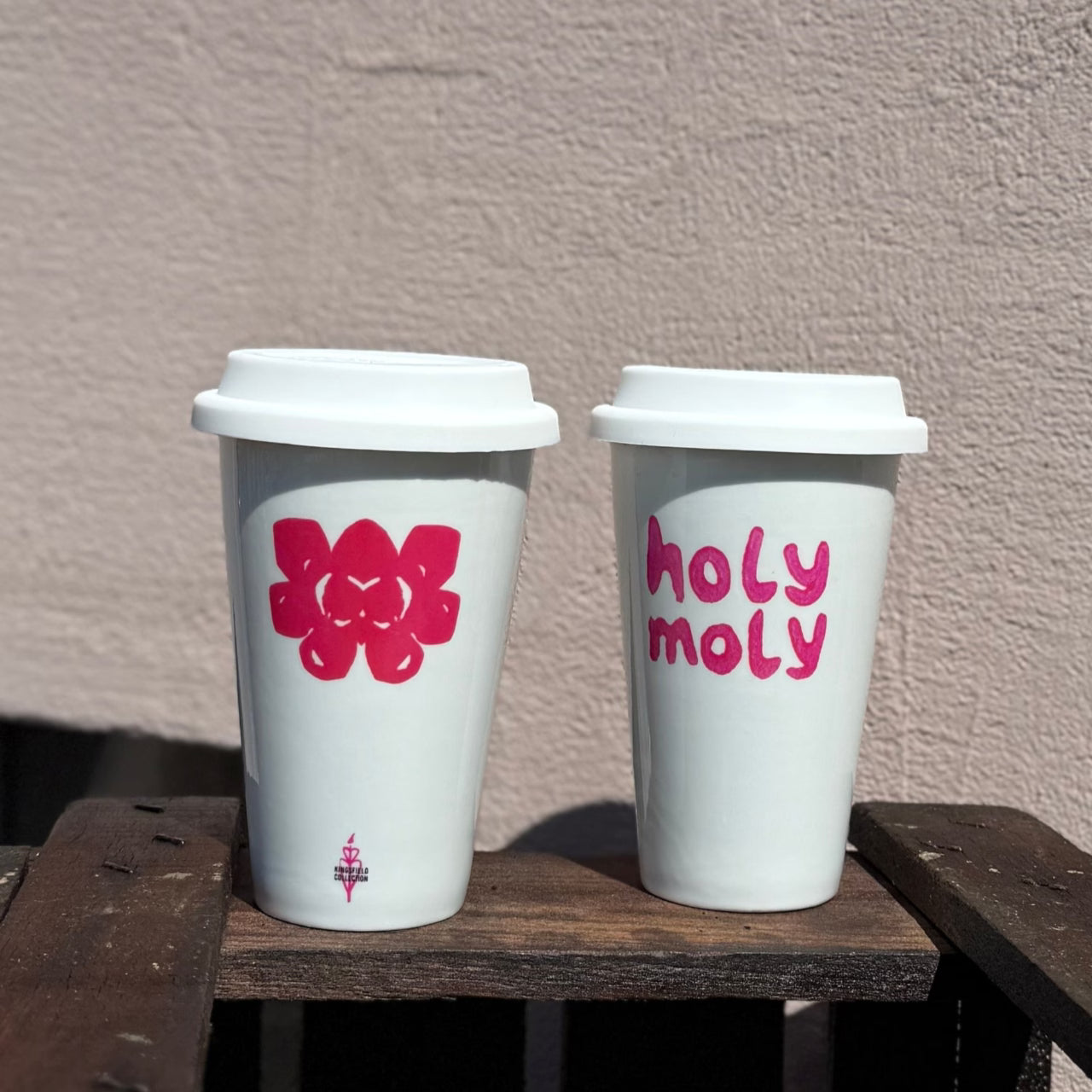 Zwei weiße To-Go-Becher mit pinkem Blume und holy moly Print stehen auf Holzkiste vor Hauswand