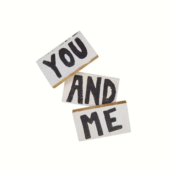 Drei Holztäfelchen vor weißem Hintergrund; jeweils schwarz auf weißer Print YOU, AND und ME